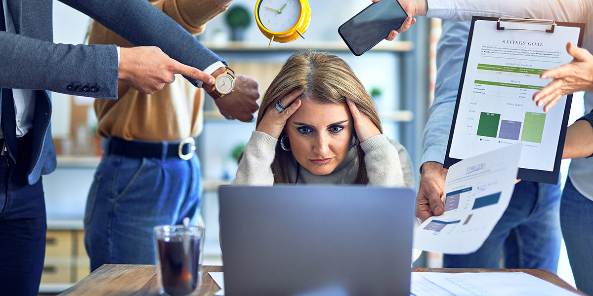 stressed woman at work surrounded by demanding coworkers knowing what causes alcoholism
