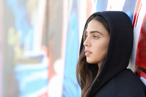 Relaxed young woman wearing a black hoodie and leaning against a wall of graffiti