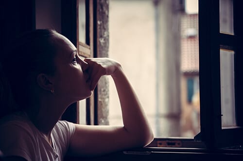 Women with hand to face, looking out window from the dark, experiencing heroin withdrawal symptoms.