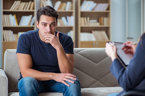 Man on couch considering info about drug addiction rehabilitation from counselor.