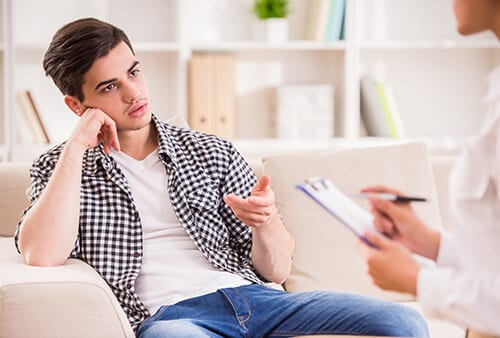 Young man on sofa talking to counselor during drug addiction therapy.