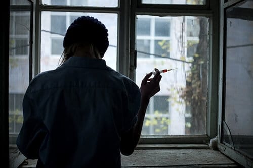 Woman in dark looking out window with needle in hand knows the heroin street names